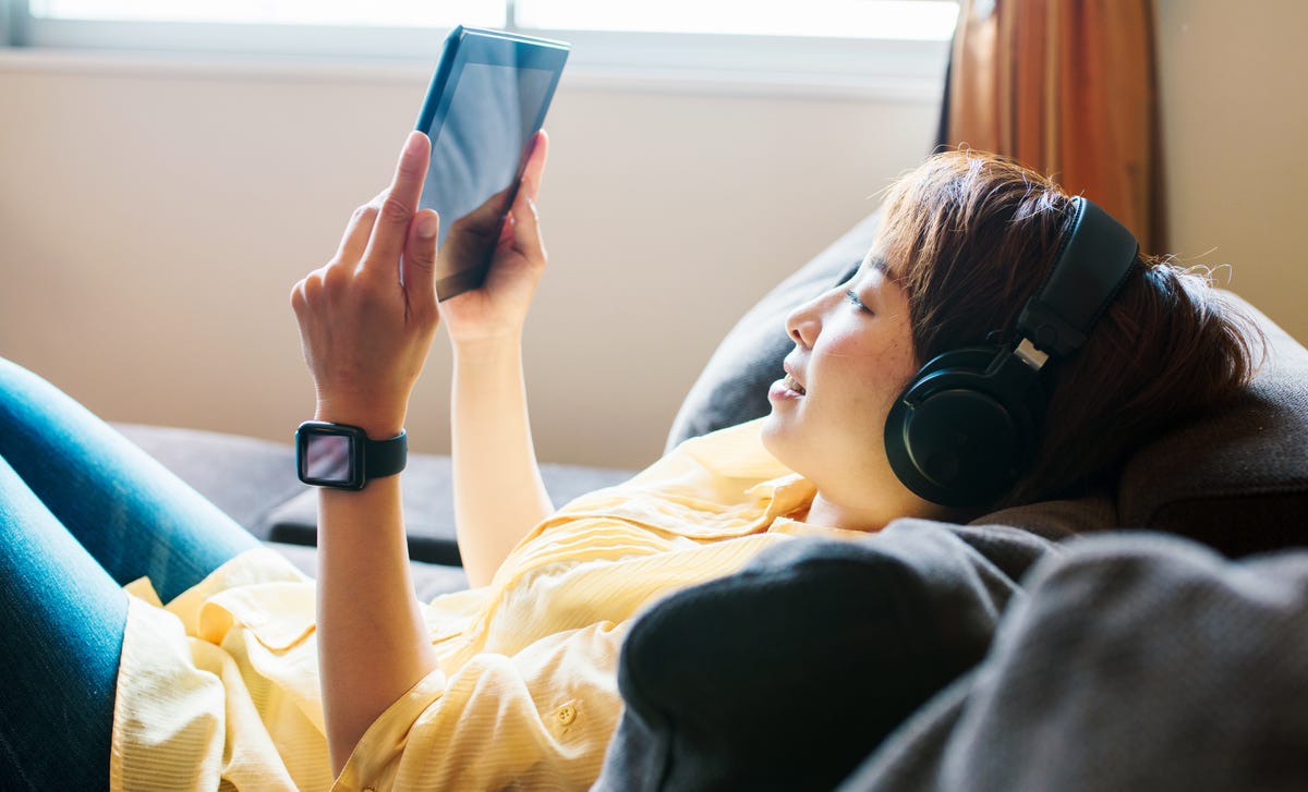 A woman relaxing on a couch, wearing headphones and using a tablet to learn Mandarin online in a comfortable home setting.