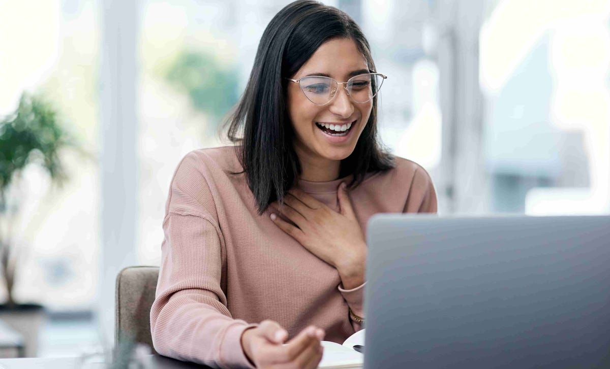A woman laughs joyfully during a video call, hand on chest, seated at a desk with a laptop.