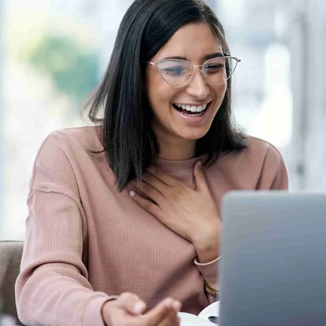 A woman laughs joyfully during a video call, hand on chest, seated at a desk with a laptop.