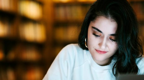 A young woman writing in a notebook while studying Urdu online in a library with a laptop.