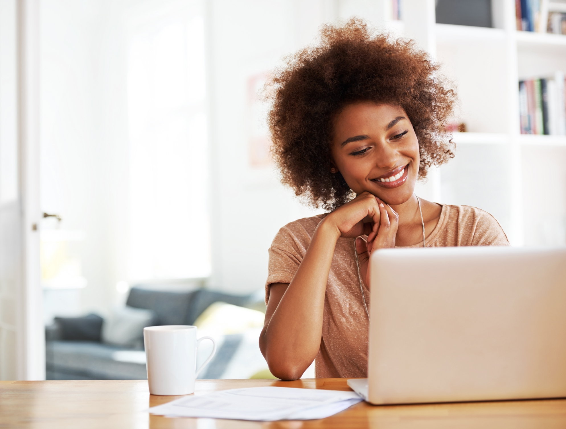 Woman smiling at her laptop while attending Italian classes online from a cozy home setup with a mug and papers on the desk.