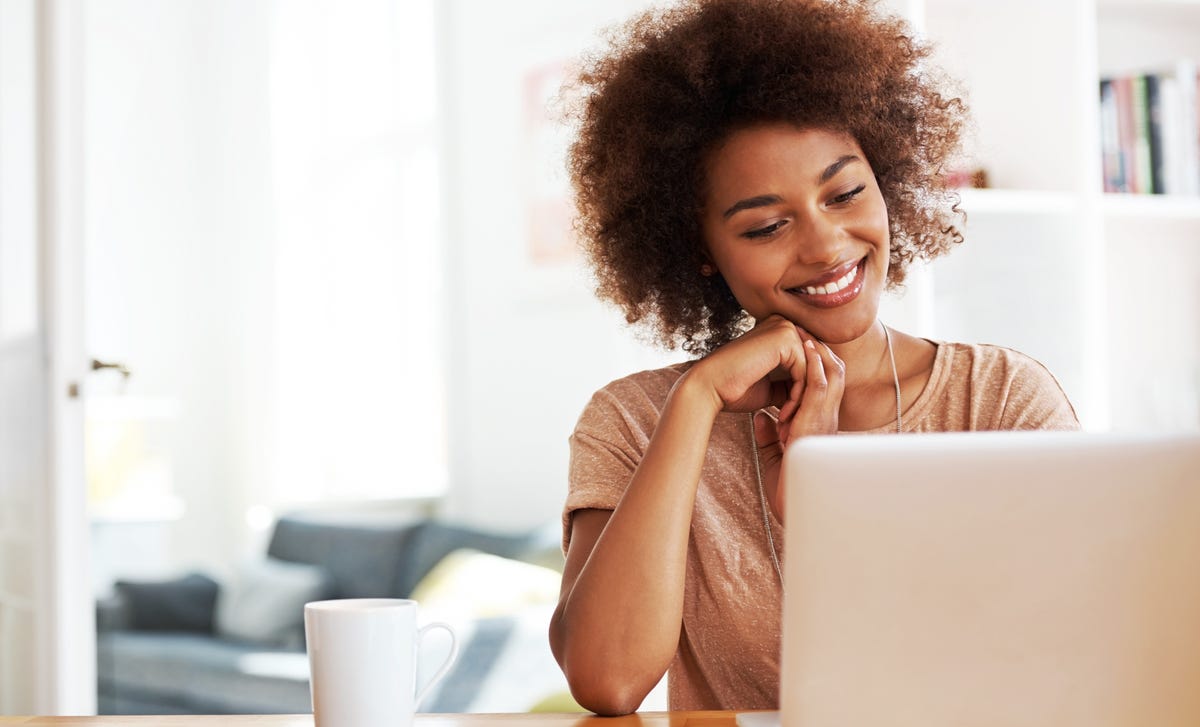 Woman smiling at her laptop while attending Italian classes online from a cozy home setup with a mug and papers on the desk.