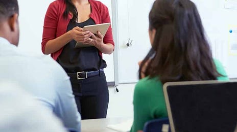 A teacher stands confidently at the front of the class, leading a language lesson using a tablet.