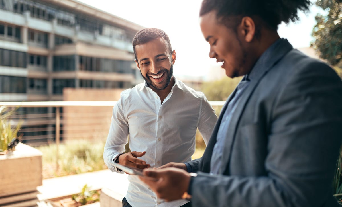 Two professional men smiling while reviewing content on a tablet, possibly discussing Turkish classes online for business communication