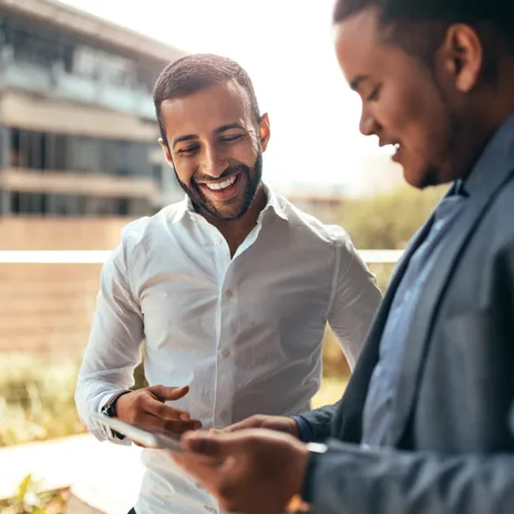 Two professional men smiling while reviewing content on a tablet, possibly discussing Turkish classes online for business communication