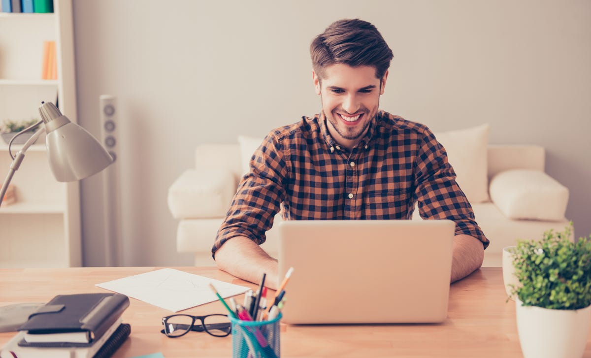 A young man smiling while studying on his laptop at home, representing an enjoyable way to learn Serbian online from a comfortable environment.