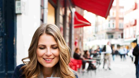 Woman smiling while using her smartphone at a café, representing the convenience of Berlitz online language schools for mobile learners.
