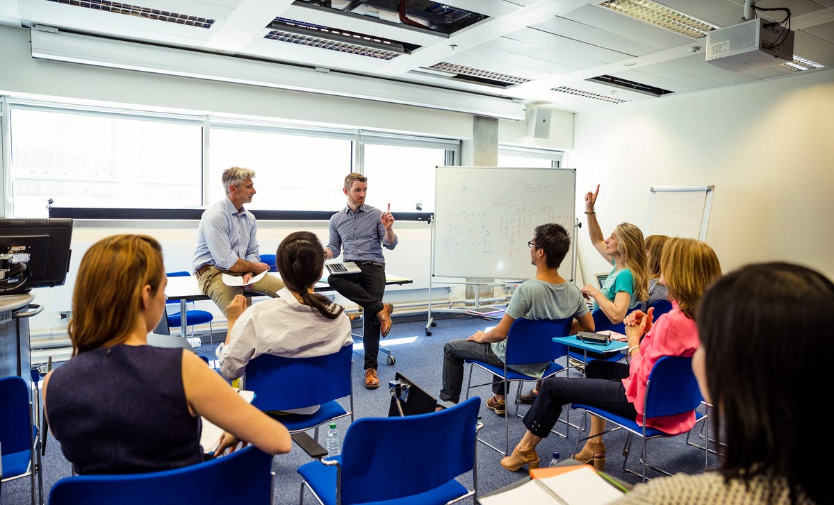 Students participating in an in-person Korean language class with instructors at the front of a modern classroom and a whiteboard filled with notes.