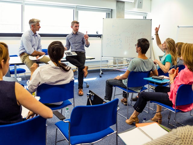 Students participating in an in-person Korean language class with instructors at the front of a modern classroom and a whiteboard filled with notes.
