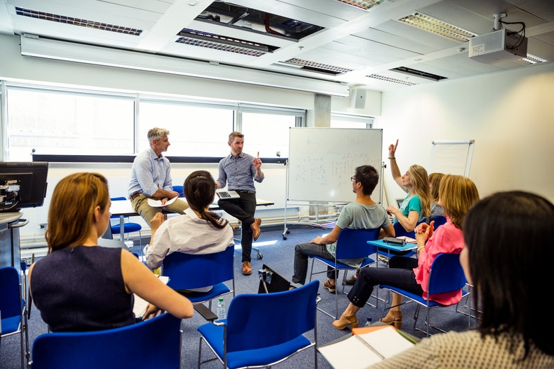 Students participating in an in-person Korean language class with instructors at the front of a modern classroom and a whiteboard filled with notes.