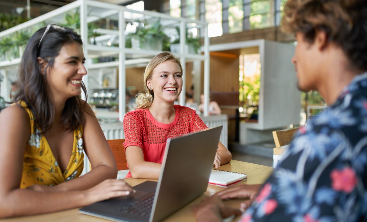 Three young adults smiling while using a laptop together at a café, appearing to learn Malaysian in a casual group setting.