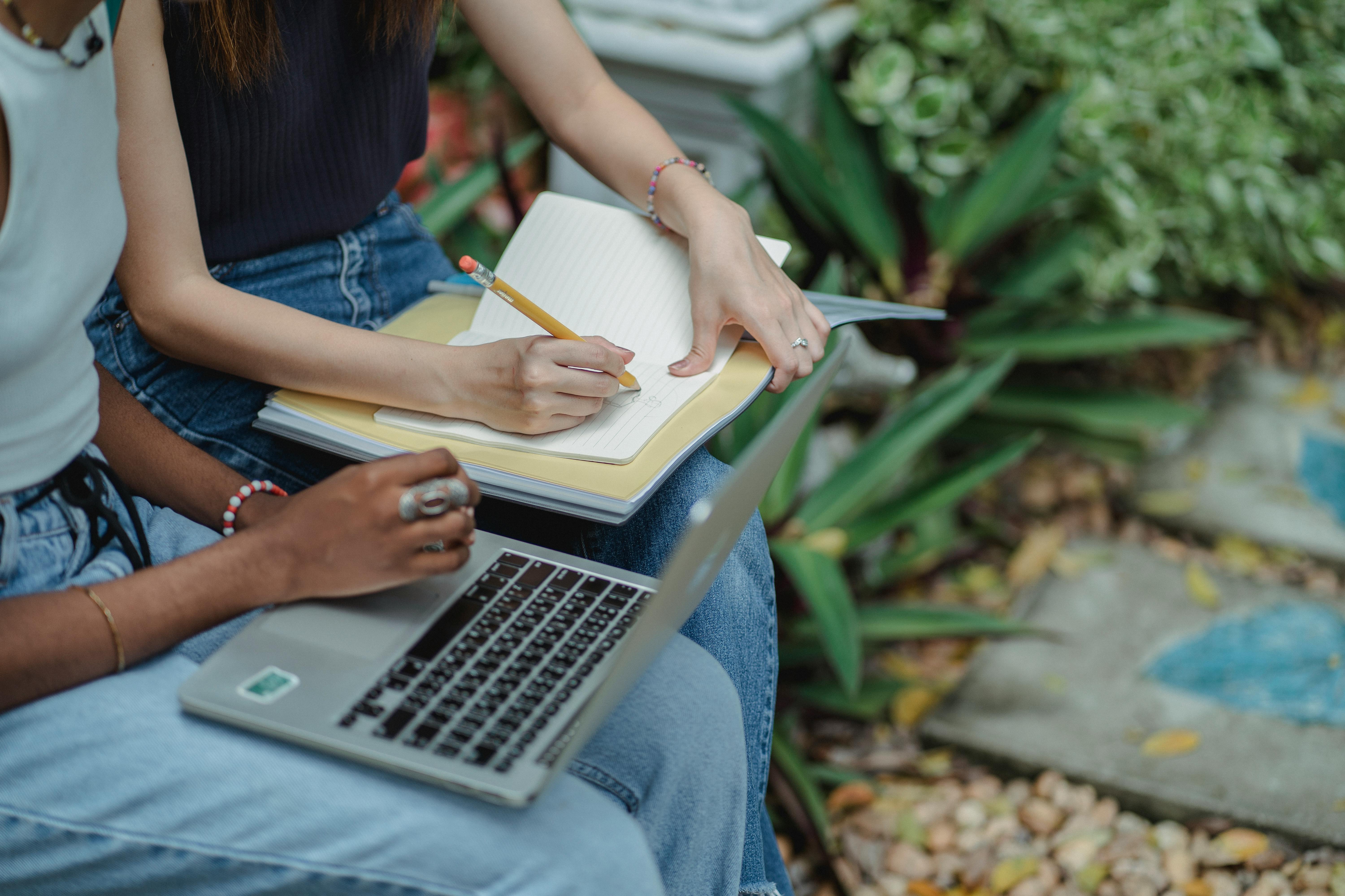 Two women sitting on a bench with their papers and laptops and practicing future continuous together