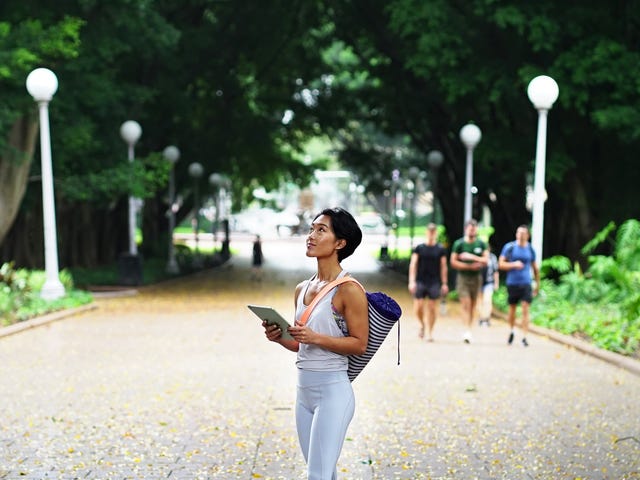 Woman holding a tablet while walking through a tree-lined park, representing mobile learning for online French classes.