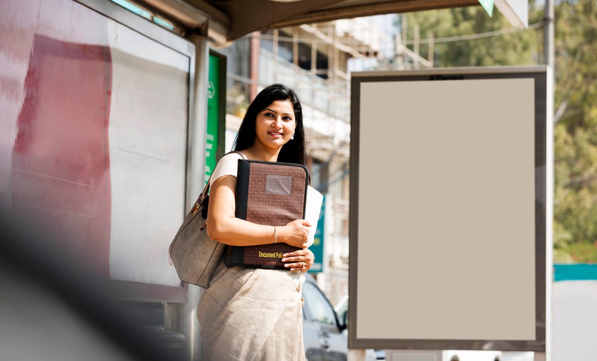 A woman standing at a bus stop holding a document folder, ready to begin her journey learning Kannada.