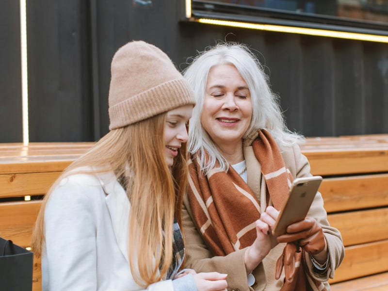 Two women sitting on a bench looking at a phone and learning about present perfect together