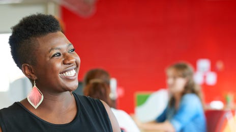 A woman with a bright smile holds a tablet in a modern, collaborative workspace, representing the accessibility and engagement of learning Haitian Creole online.