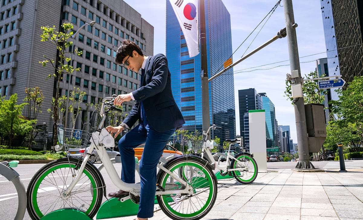 Man in business attire unlocking a bike at a public station in South Korea, with a Korean flag and city buildings in the background—symbolizing everyday life and Korean language immersion.