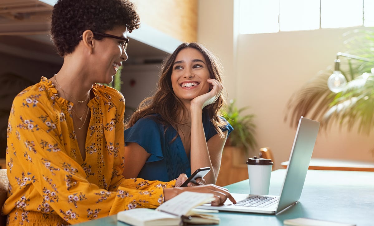 Two smiling women study together with a laptop and smartphone, representing the joy and convenience of learning Turkish online in a modern, bright setting.