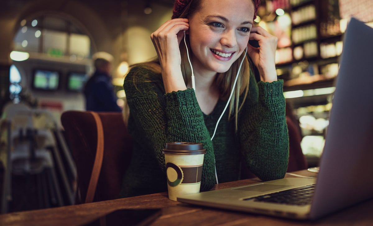 A young woman wearing a green sweater and red hat smiles while learning Swedish online on her laptop in a coffee shop, enjoying a warm beverage.