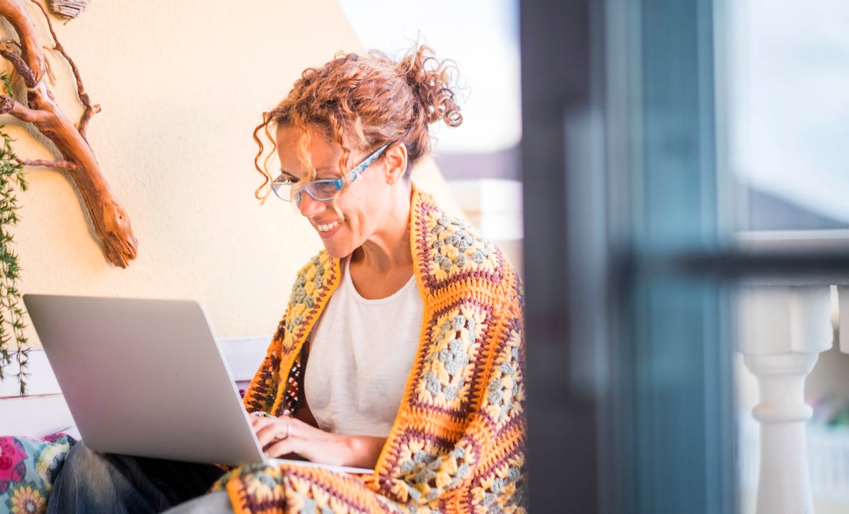 A smiling woman wrapped in a colorful blanket studies with her laptop, representing the comfort and flexibility of learning Hindi online from home.