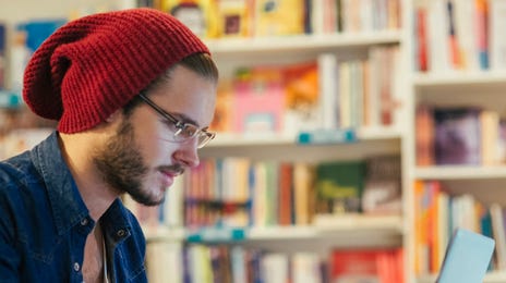 A young man wearing a red beanie studies with his laptop and coffee in a modern library, representing the casual and comfortable approach to learning Danish online.