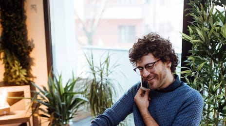 A young man smiles while working on his laptop in a bright home office, representing the accessibility and convenience of learning Turkish online from home.