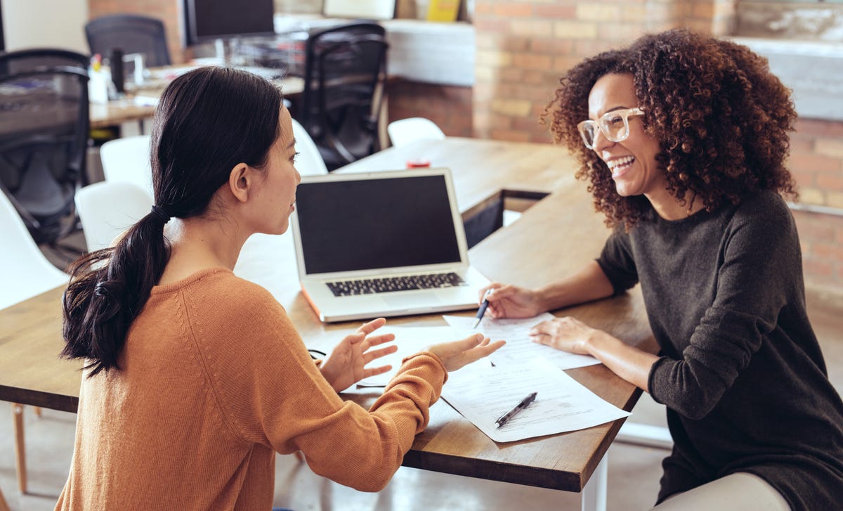 Two women in a classroom setting smiling and talking while reviewing notes during an in-person French class session