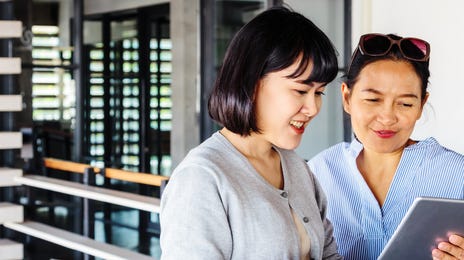 Two women smiling while using a tablet outside a modern building, symbolizing a collaborative and enjoyable way to learn Malaysian online.