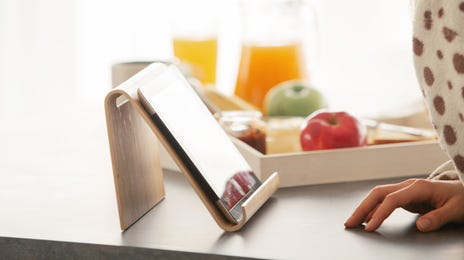 A woman in a cozy polka-dot robe smiles at a tablet on a kitchen counter, enjoying a relaxed breakfast while she learns Swedish online.