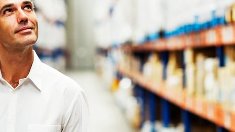 Middle-aged man in a white shirt standing in a warehouse aisle, thoughtfully looking up—symbolizing reflection and motivation to learn Korean.