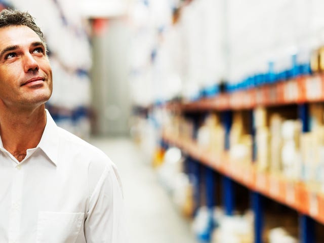 Middle-aged man in a white shirt standing in a warehouse aisle, thoughtfully looking up—symbolizing reflection and motivation to learn Korean.