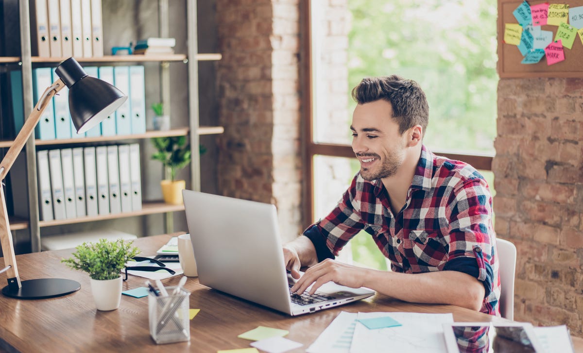 A young man in a plaid shirt smiling while studying Indonesian online on his laptop in a bright, modern office.
