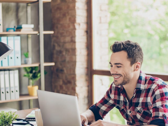 A young man in a plaid shirt smiling while studying Indonesian online on his laptop in a bright, modern office.