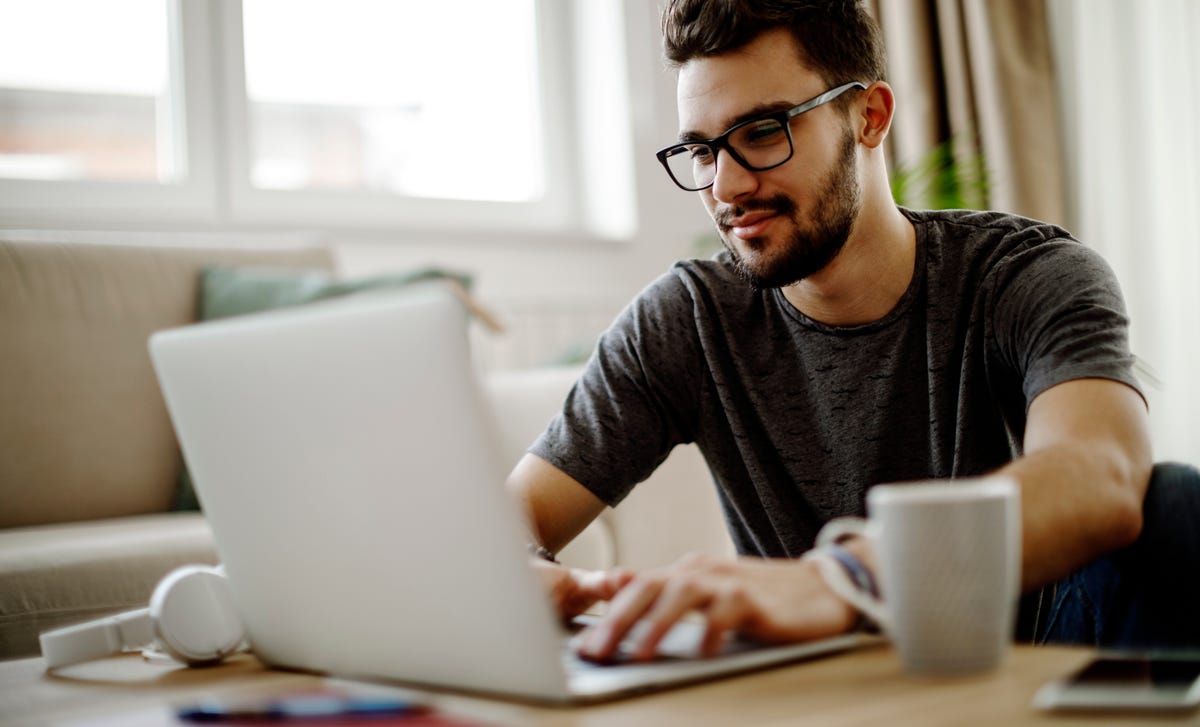 A young man in a casual t-shirt uses his laptop to learn Urdu online at home with a coffee cup nearby, showcasing a relaxed study atmosphere.