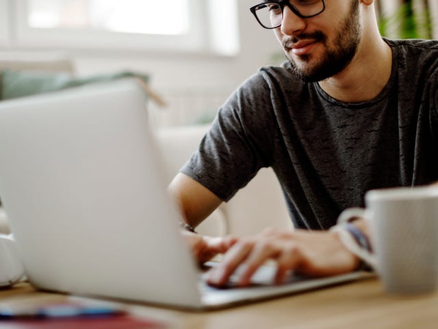 A young man in a casual t-shirt uses his laptop to learn Urdu online at home with a coffee cup nearby, showcasing a relaxed study atmosphere.