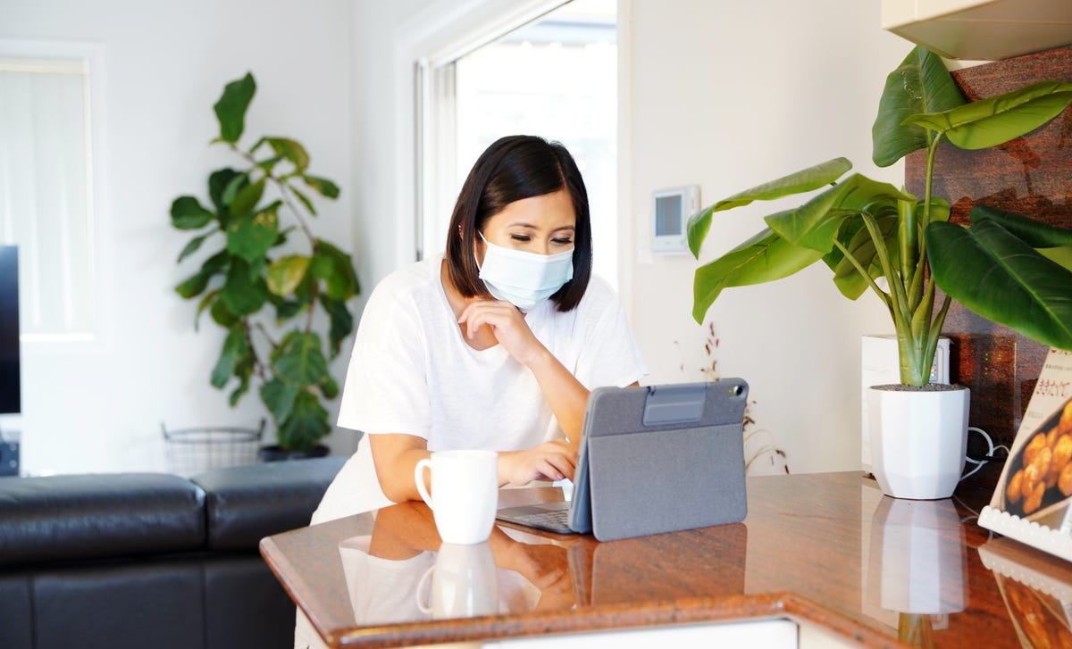 A young woman wearing a mask studies on a tablet from home, representing the concept of learning Vietnamese online in a comfortable environment.