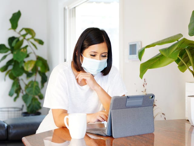 A young woman wearing a mask studies on a tablet from home, representing the concept of learning Vietnamese online in a comfortable environment.