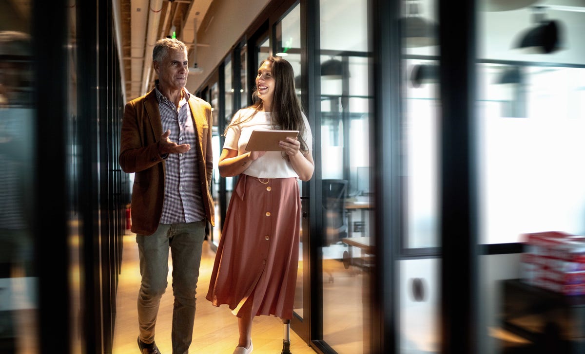 Two colleagues in a modern office hallway discussing strategies for a German teaching career, with a young woman holding a tablet and wearing a prosthetic leg.