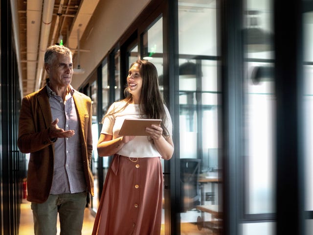 Two colleagues in a modern office hallway discussing strategies for a German teaching career, with a young woman holding a tablet and wearing a prosthetic leg.