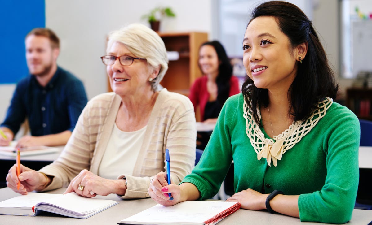 Adults attentively participating in a Finnish language course in a classroom setting, taking notes and listening to the instructor
