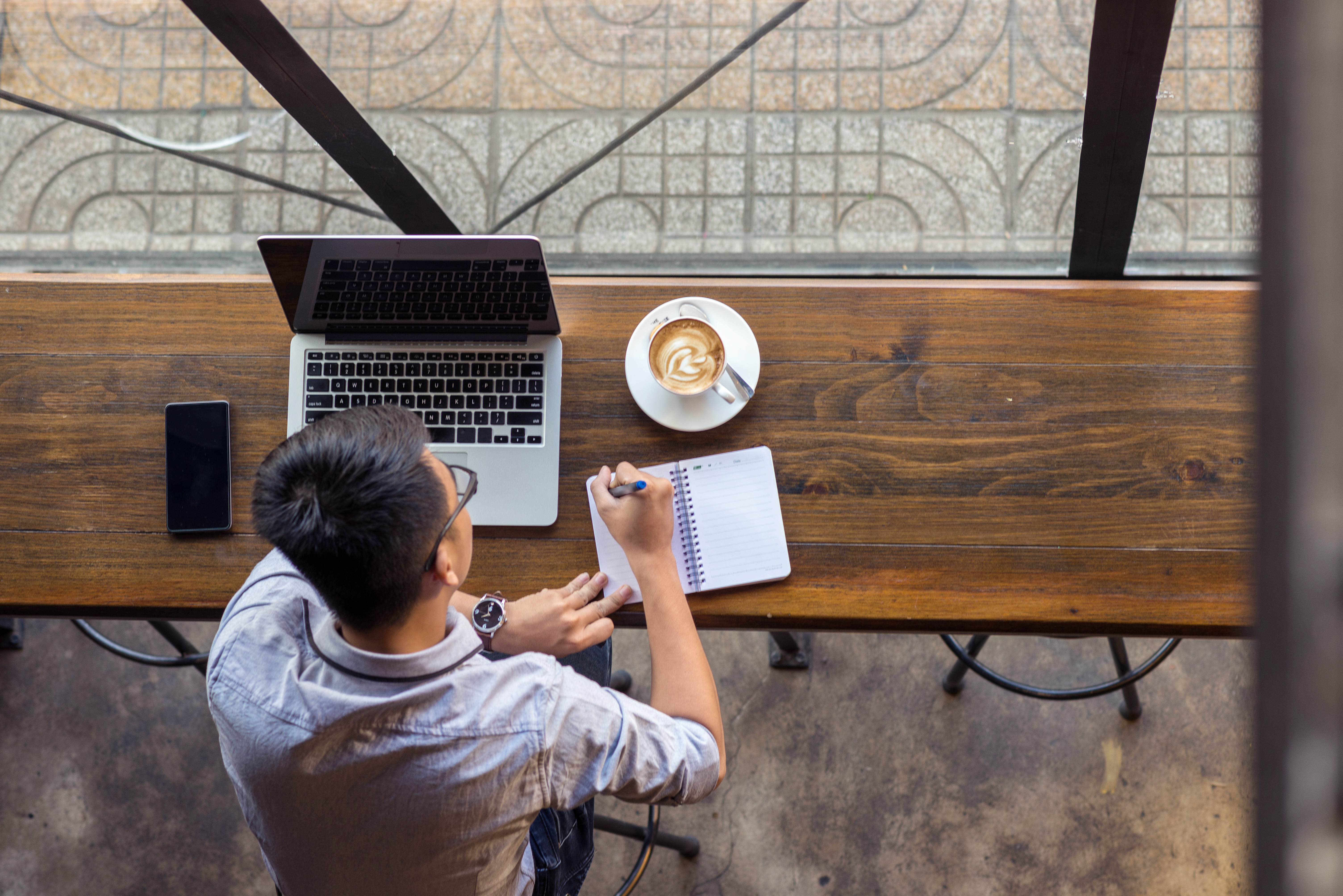 Top view of a man studying Korean online in a coffee shop with a laptop, notebook, and latte on a wooden table.