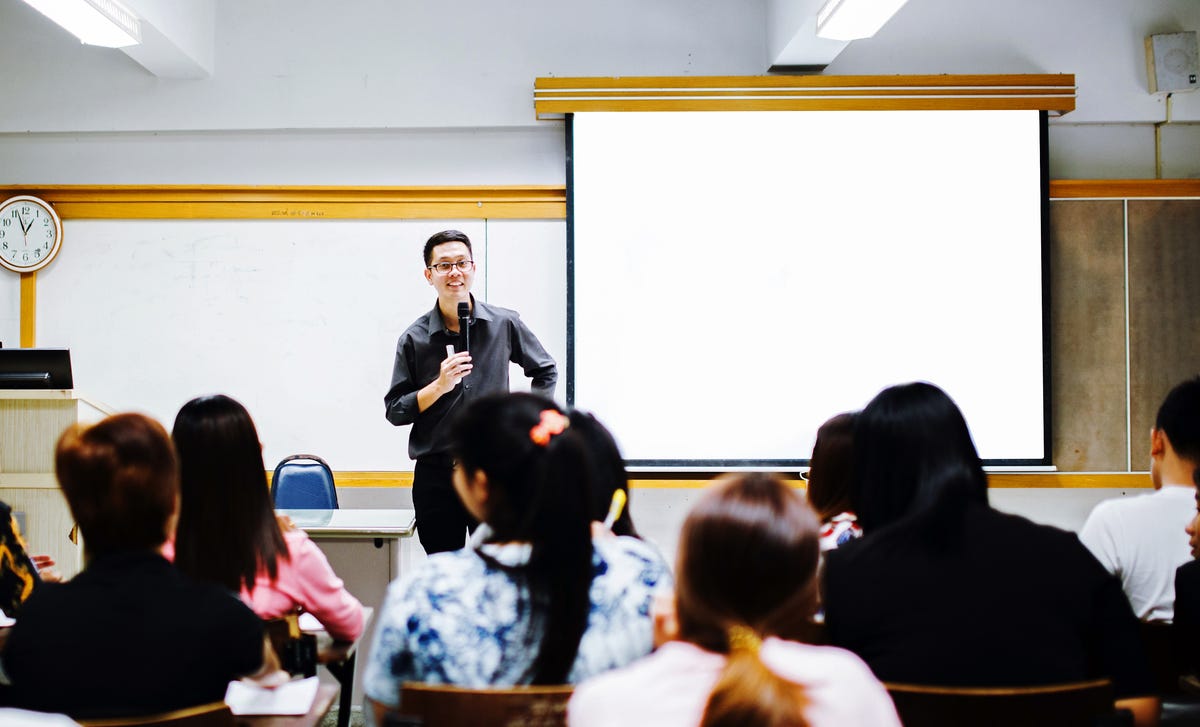 A teacher holds a microphone and smiles while conducting Indonesian language lessons in a classroom with students paying close attention.