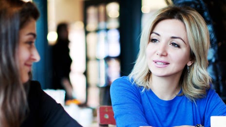 Two women engaged in a lively conversation at a colorful café, illustrating the interactive and social way to learn Latvian through conversation and real-world practice.