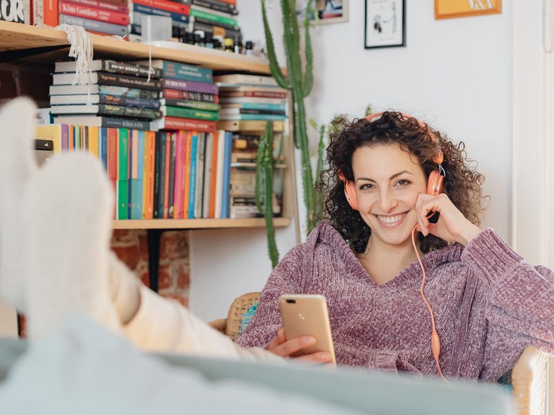 A smiling woman with her feet up is learning a language through music at home with headphones,