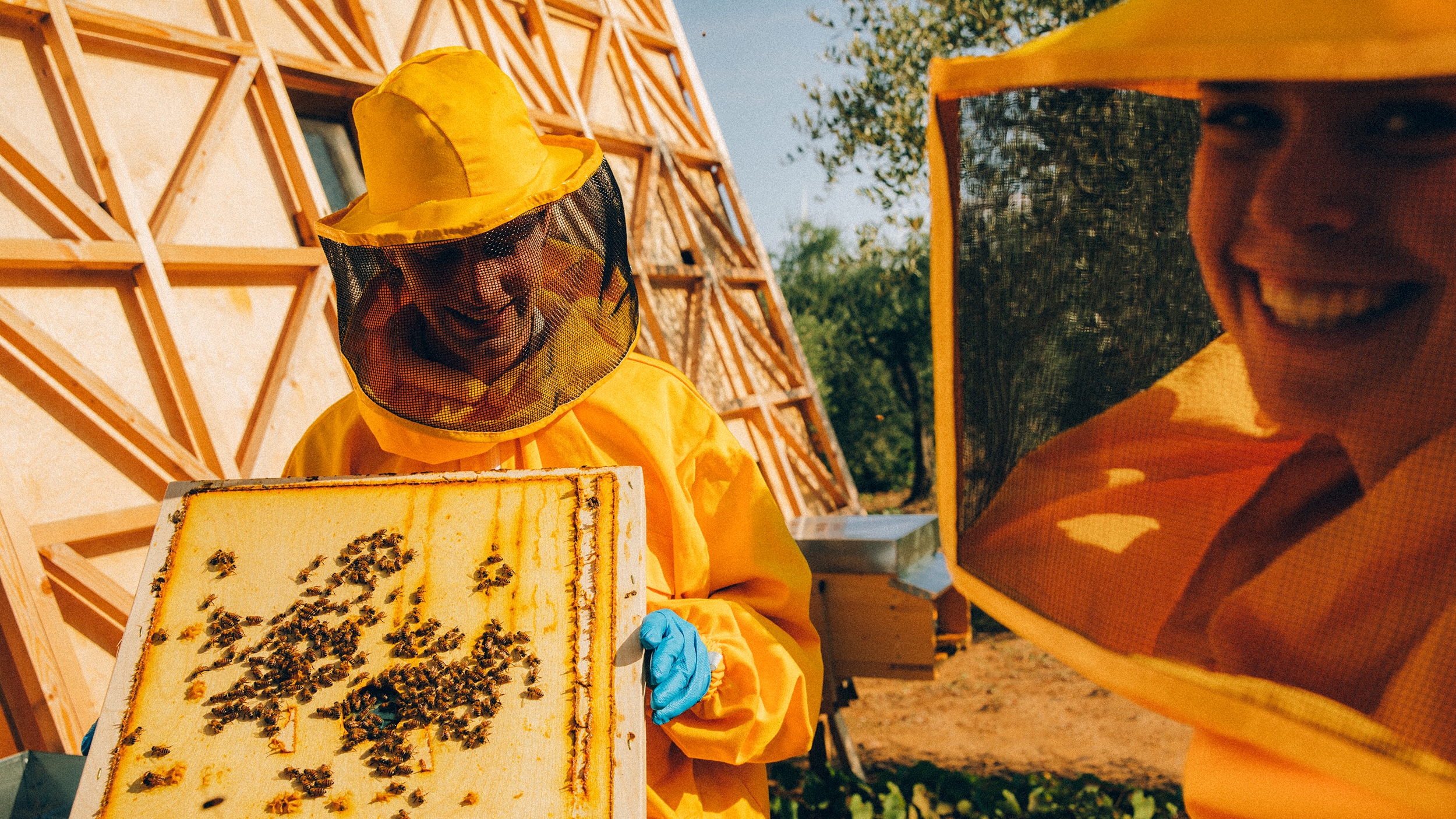 Dos personas con trajes de apicultor de color naranja brillante sonríen bajo el sol, de pie sobre una tabla de madera salpicada de abejas.