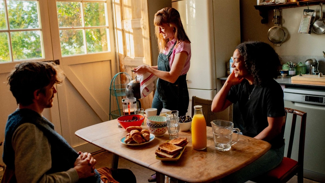 Drie Airbnb-gasten aan een keukentafel met gebakjes, geroosterd brood en sap. Eentje maakt koffie in een Franse pers.