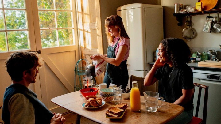 Tres huéspedes en Airbnb se reúnen alrededor de una mesa de cocina con tortas, tostadas y jugos. Uno prepara café en una prensa francesa.