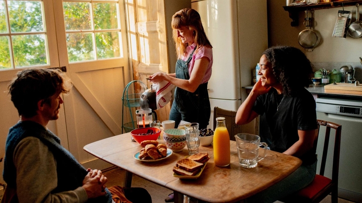 Three Airbnb guests gather at a kitchen table topped with pastries, toast, and juice. One is making coffee in a French press.