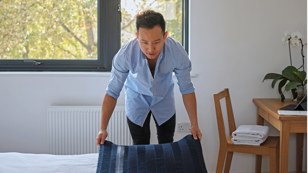 一個穿著帶鈕扣的藍色襯衫的人把毯子鋪在床尾，旁邊的桌子上放著一盆白蘭花。