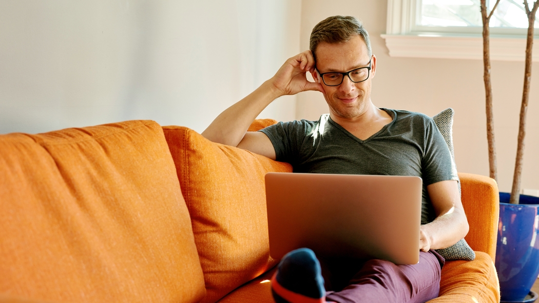 Un hombre con lentes está sentado en un sofá naranja con una laptop en sus piernas.